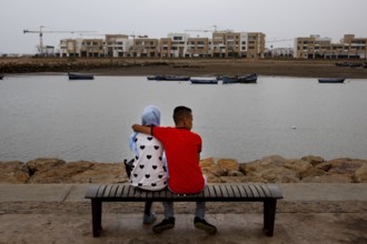 A couple sits on a bench on the riverbank with a view of modern buildings and cranes, Rabat, Salé,