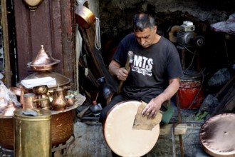 Craftsman repairs copper dishes surrounded by traditional tools and metalwork, Fez, Fez el Bali,