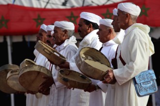 Men wearing traditional clothes holding drums in front of a backdrop with Berber decoration, El