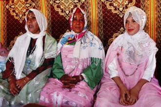 Three woman in traditional dress sit in a tent during the rose festival in El Kelâa m'Gouna, El