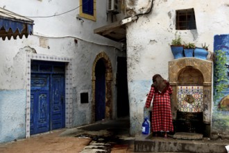 A woman draws water from a spring next to a blue gate in the old town, Essaouira, Morocco