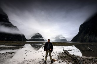 Man standing in front of a dramatic sky on the shore of a fjord with reflecting water, Milford