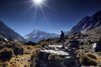 A hiker stands on a rocky trail with views of mountain peaks in sunshine, Mount Cook, New Zealand