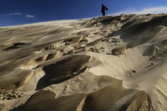 Person climbing large sand dunes during a sandstorm under a blue sky