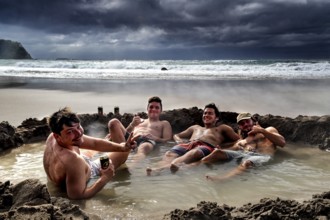 Group of men relaxing in waterhole on beach under dramatic clouds, Hot Water Beach, Coromandel