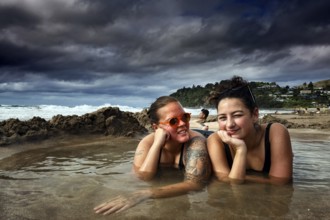 Two woman lying relaxing in a puddle of water on a beach under cloudy sky, Hot Water Beach,