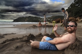 Man relaxing in sand pit on cloudy Hot Water Beach, Coromandel Peninsula, Waikato, New Zealand