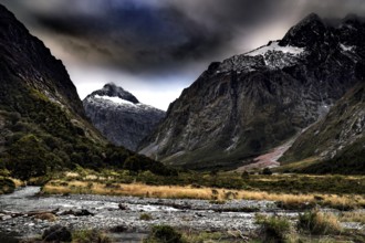 Dramatic landscape with snow-capped mountains and a river in the foreground under a cloudy sky,