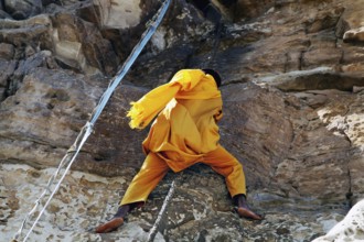 A monk dressed in yellow climbs a rock with a rope, Debre Damo, Tigray, Ethiopia