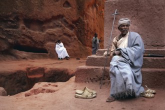 A priest with a cross sits near the rock church of Bete Emanuel in Lalibela, Lalibela, Amhara,