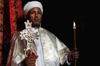 Priest in white cloak majestically highlights a cross in the dark interior of Bete Meskel,