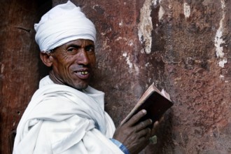 A man in traditional dress reads with a smile on a stone wall, Lalibela, Amhara, Ethiopia