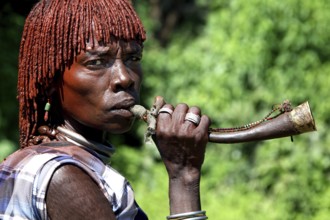 Hamer woman blowing a traditional horn surrounded by green vegetation, Turmi, Ethiopia