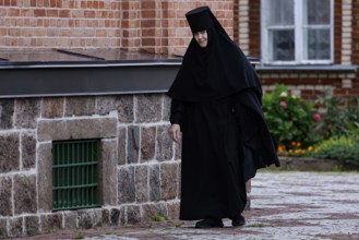 Nun in black clothes walks along the stone wall of the church, Kuremäe, Pühtitsa, Estonia