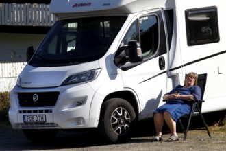 Female camper relaxing next to a white motorhome under a blue sky, Moskenesøy, Lofoten, Norway