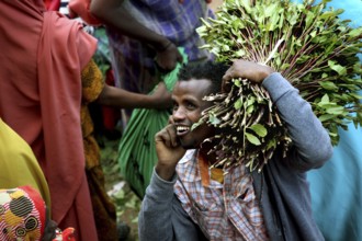 Smiling man carrying khat bundles on his shoulder and talking on the phone at a market, Awaday,