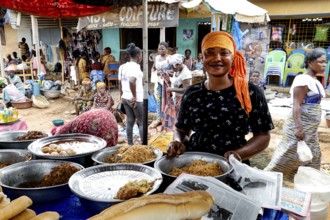 Woman in a busy market smiling behind a table with various foods, Brobo, Côte d'Ivoire