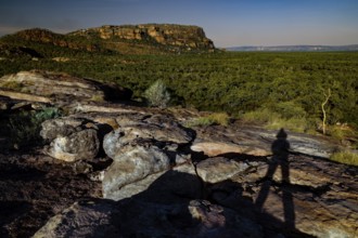 Shadows of a person on a rocky elevation in front of forest and rocks under clear sky, Nawurlandia,