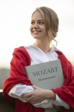 Smiling young woman, pianist, in white blouse and red cardigan, in the evening light, holding notes