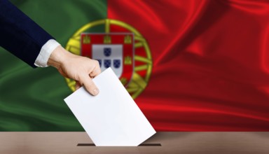 Hand placing a ballot in ballot box with the Portuguese flag in the background. Portugal