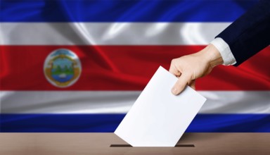 Hand placing a ballot in ballot box with the Costa Rican flag in the background. Costa Rica