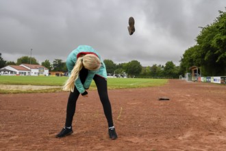 A woman on a sports field throws a rubber boot into the air under a cloudy sky, wellies long throw