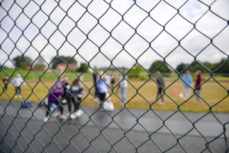 Photographed through a wire fence, a blurred group of spectators is seen on a sports field, Lower
