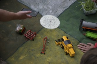 Hand points at a miniature agricultural model with toy vehicles on a table, event for