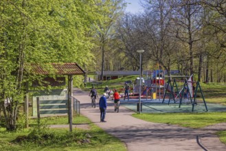 People cycling and walking by a playground with swings on a sunny spring day in a public park,