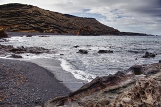 Secluded beach with black sand surrounded by wavy rock formations under a cloudy sky, Chargo Verde