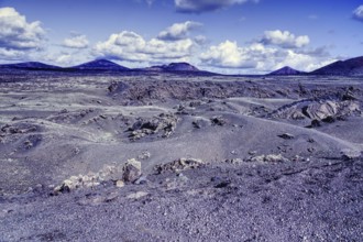 Wide, dry volcanic landscape with rolling hills and mountains under a cloudy blue sky, Tinajo