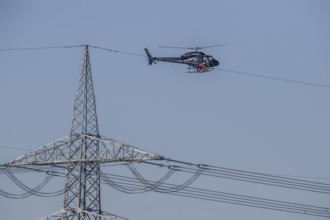 A helicopter carries out technical work on power lines next to a mast under a blue sky, an employee