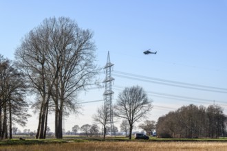 A helicopter flies over a landscape with power poles and trees under a blue sky, Lemförde, Germany,