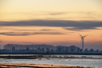 A wind turbine at sunset next to a calm body of water and trees, wind turbines in front of an