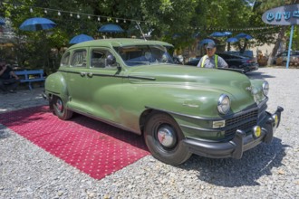 Green vintage car standing on outdoor red carpet surrounded by colorful umbrellas and gravel floor,