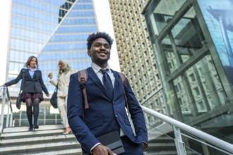 Confident black businessman walking down modern city stairs, carrying a tablet and backpack, with