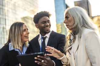 Diverse business professionals meet outdoors in the city, smiling and collaborating over a tablet