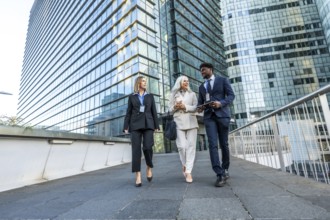 Three multi ethnic business colleagues walking and talking on a pedestrian bridge amid modern glass