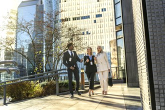 Three diverse business professionals, a black man and two white women, walking outdoors on a sunny