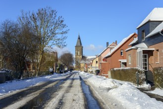 Wintery village road with snow cover, surrounded by houses and a church tower under a blue sky,