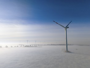 Wind turbine rises above foggy snow-covered landscape in clear blue sky, aerial view, wind turbine