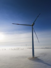 A wind turbine stands out in a snowy, foggy landscape, surrounded by soft morning light and clear