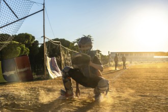 Boy catcher wearing protective gear and mitt, squatting on a dusty baseball field with golden