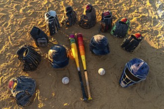 Baseball bats, gloves, helmets and catcher's mask arranged in a circle on a sandy field at sunset,