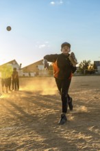 Young boy in baseball uniform pitching a baseball, creating a cloud of dust on the field during an