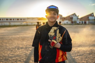 Young male baseball player stands confidently on a sunlit dirt field at golden hour, cap on and