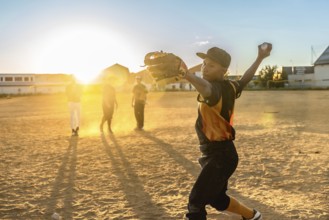 Boy pitcher throws a baseball on a dusty summer field at golden hour, practicing with teammates,