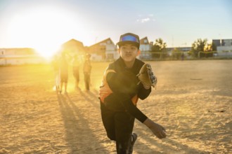 Young boy in baseball uniform and glove pitching a baseball on a dirt field during sunset, with