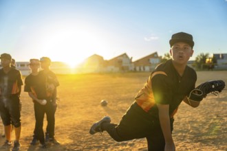 Young baseball pitcher in uniform and cap winds up to throw on a dusty field at golden sunset,
