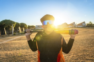 Youth baseball player in sunglasses and orange black jersey gripping a bat on a dusty field at